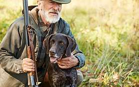 Ein älterer Mann mit einem Hund und einem Gewehr in der Natur.